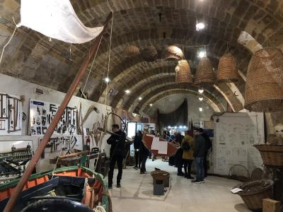 The image shows the interior of a museum with stone vaulted ceilings, featuring exhibits of traditional fishing boats, large woven fish traps (known as "trappole" or "nasse"), and informational displays about fishing tools and methods.