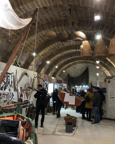 The image shows the interior of a museum with stone vaulted ceilings, featuring exhibits of traditional fishing boats, large woven fish traps (known as "trappole" or "nasse"), and informational displays about fishing tools and methods.