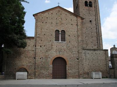 A low-angle photograph of a large, ancient brick basilica with a prominent square bell tower, featuring arched windows, a central wooden door, and two stone sarcophagi in the foreground plaza.