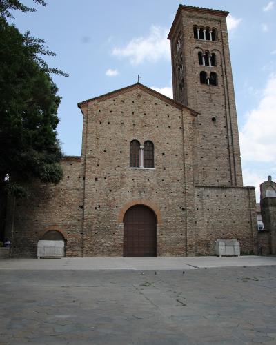A low-angle photograph of a large, ancient brick basilica with a prominent square bell tower, featuring arched windows, a central wooden door, and two stone sarcophagi in the foreground plaza.
