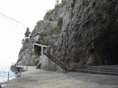 A coastal cliffside scene showing a paved walkway, stone stairs leading up a rocky face, and the dark entrance to a sea cave on the right, with the sea visible on the left.