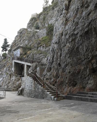 A coastal cliffside scene showing a paved walkway, stone stairs leading up a rocky face, and the dark entrance to a sea cave on the right, with the sea visible on the left.