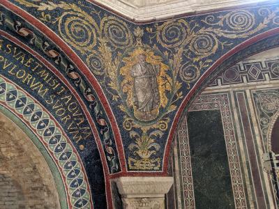 The image shows an interior view of a historic religious structure featuring elaborate Byzantine-style mosaics on vaulted ceilings and arches, with Latin inscriptions and a central figure in a medallion surrounded by golden foliage; stone columns