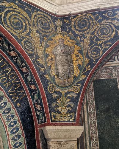 The image shows an interior view of a historic religious structure featuring elaborate Byzantine-style mosaics on vaulted ceilings and arches, with Latin inscriptions and a central figure in a medallion surrounded by golden foliage; stone columns