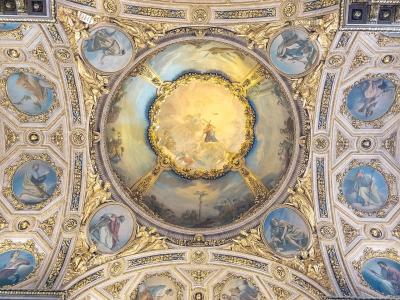 The image shows an ornate, gilded Baroque ceiling with a central fresco depicting a religious scene surrounded by smaller oval paintings of saints or biblical figures, framed by elaborate stucco and gold leaf decorations.