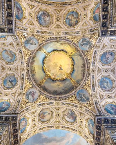 The image shows an ornate, gilded Baroque ceiling with a central fresco depicting a religious scene surrounded by smaller oval paintings of saints or biblical figures, framed by elaborate stucco and gold leaf decorations.
