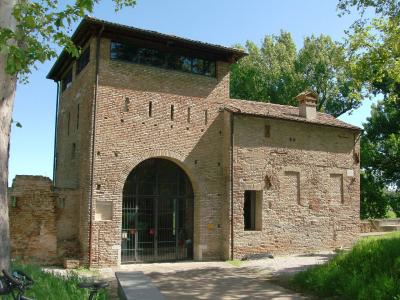 A two-story brick building with an arched gateway on the ground floor, featuring a tower-like upper section with windows and a tiled roof, set against green trees under a clear blue sky.