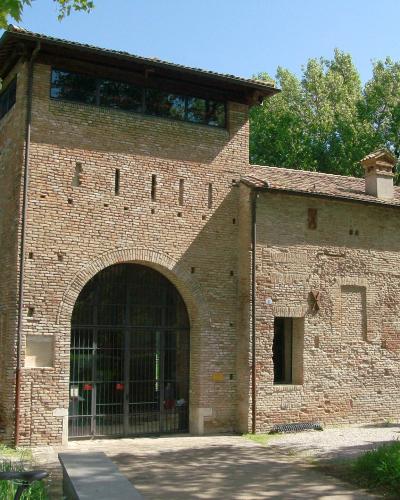 A two-story brick building with an arched gateway on the ground floor, featuring a tower-like upper section with windows and a tiled roof, set against green trees under a clear blue sky.