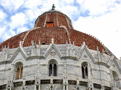 A low-angle view of the ornate, terracotta-tiled dome and upper facade of a large Romanesque building with white marble detailing, arched windows, and decorative statues, set against a cloudy sky.