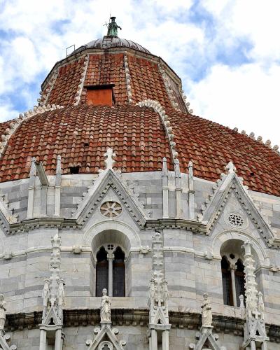 A low-angle view of the ornate, terracotta-tiled dome and upper facade of a large Romanesque building with white marble detailing, arched windows, and decorative statues, set against a cloudy sky.