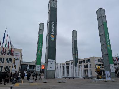 The image shows the exterior of Rimini Fiera, featuring its distinctive tall glass towers with "Iberdrola" branding, fountains in the plaza, and signage indicating "Padiglione B3 Stand 101," with people walking nearby under an overcast sky.