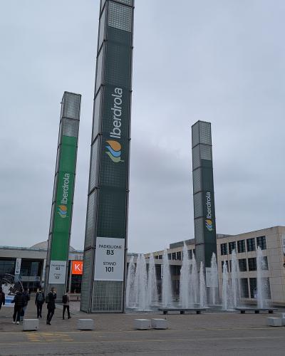 The image shows the exterior of Rimini Fiera, featuring its distinctive tall glass towers with "Iberdrola" branding, fountains in the plaza, and signage indicating "Padiglione B3 Stand 101," with people walking nearby under an overcast sky.