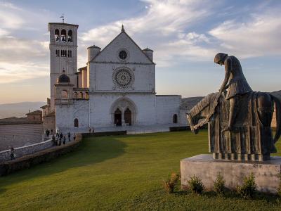 The image shows the Basilica of San Francesco in Assisi, Italy, featuring its distinctive white stone facade, rose window, bell tower, and a modern bronze equestrian statue in the foreground.