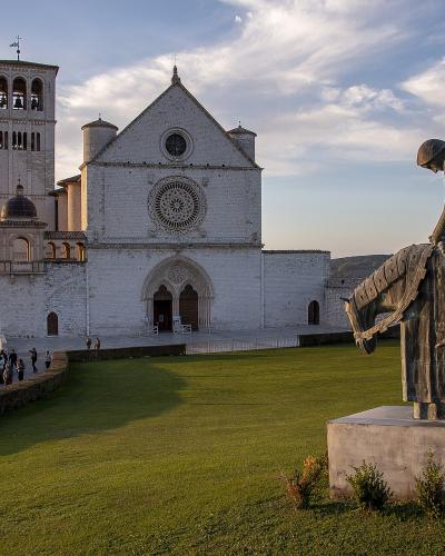 The image shows the Basilica of San Francesco in Assisi, Italy, featuring its distinctive white stone facade, rose window, bell tower, and a modern bronze equestrian statue in the foreground.