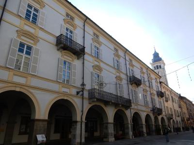 A low-angle view of a long, pale yellow building with a continuous ground-floor arcade of rounded arches, upper-story windows with white shutters and wrought-iron balconies, and a clock tower visible in the background under a bright sky.