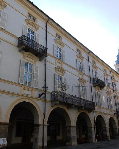 A low-angle view of a long, pale yellow building with a continuous ground-floor arcade of rounded arches, upper-story windows with white shutters and wrought-iron balconies, and a clock tower visible in the background under a bright sky.