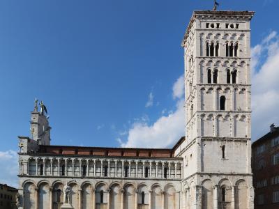 The image shows the exterior of a Romanesque-style church with a prominent tall bell tower (campanile) and a facade featuring multiple tiers of blind arches, set in a paved public square under a blue sky; the filename explicitly identifies it as '