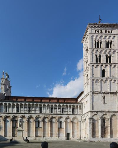 The image shows the exterior of a Romanesque-style church with a prominent tall bell tower (campanile) and a facade featuring multiple tiers of blind arches, set in a paved public square under a blue sky; the filename explicitly identifies it as '