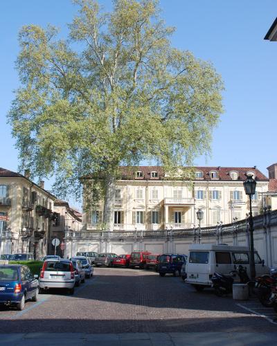 The image shows a large, pale yellow neoclassical building with a red-tiled roof and a prominent balcony, partially obscured by a tall, leafy tree, situated behind a low stone wall and a cobblestone parking area with several cars and scooters.