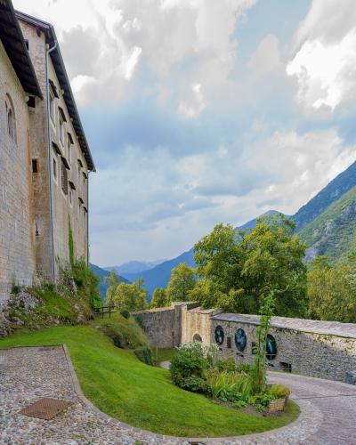 The image shows the exterior stone walls of a historic castle complex with a grassy slope, a paved path, and a lower stone wall featuring circular decorative elements, set against a backdrop of green mountains under a cloudy sky.