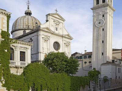 A white Baroque-style church with a prominent dome and a tall, slender bell tower (campanile) stands beside a canal, with lush green ivy covering a wall in the foreground.
