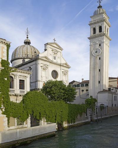 A white Baroque-style church with a prominent dome and a tall, slender bell tower (campanile) stands beside a canal, with lush green ivy covering a wall in the foreground.
