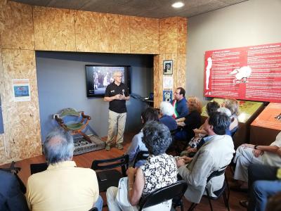 The image shows an indoor presentation or lecture taking place in a room with OSB wood paneling, where a man is speaking to an audience seated in chairs, with a red informational wall panel titled "The Mission of the Museum" visible on the right.