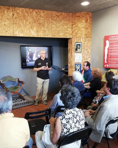 The image shows an indoor presentation or lecture taking place in a room with OSB wood paneling, where a man is speaking to an audience seated in chairs, with a red informational wall panel titled "The Mission of the Museum" visible on the right.