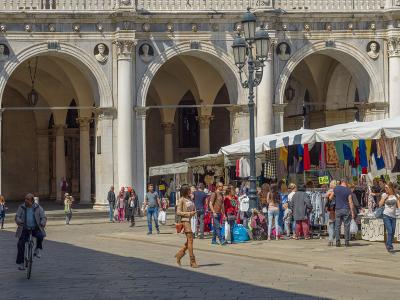 The image shows a sunlit, paved public square with a prominent Renaissance-style loggia featuring arched colonnades and sculpted medallions, alongside market stalls under white canopies selling colorful textiles and goods, with pedestrians and a c
