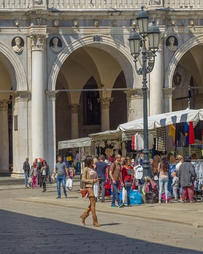 The image shows a sunlit, paved public square with a prominent Renaissance-style loggia featuring arched colonnades and sculpted medallions, alongside market stalls under white canopies selling colorful textiles and goods, with pedestrians and a c