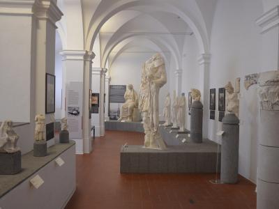 The interior of a museum gallery featuring classical marble statues, busts, and architectural fragments displayed on pedestals under white vaulted ceilings with terracotta tile flooring.