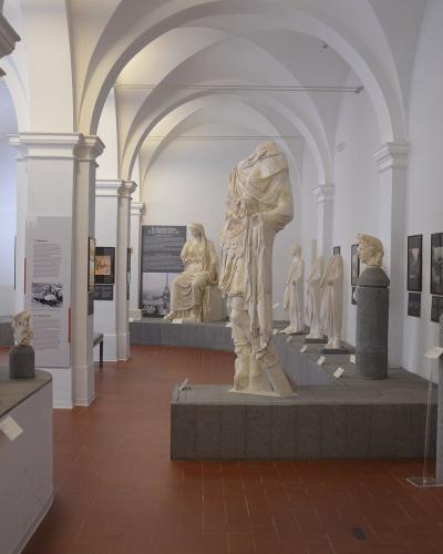 The interior of a museum gallery featuring classical marble statues, busts, and architectural fragments displayed on pedestals under white vaulted ceilings with terracotta tile flooring.