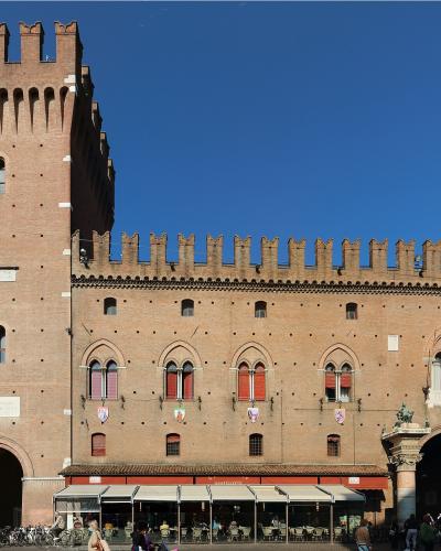The image shows a large, historic brick building with a prominent crenellated tower on the left, arched windows, and a ground-floor arcade with outdoor seating; signage includes Italian flags and coats of arms, and a statue of a figure on horsebac