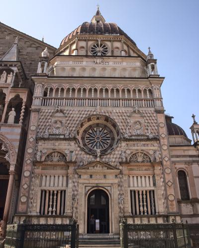 The image shows the ornate Renaissance facade of a chapel with a distinctive diamond-patterned marble exterior, a large central rose window, and a dome, situated next to a larger basilica structure under a clear blue sky.