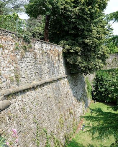 A section of a high, sloping stone fortification wall with a stone parapet and a drainpipe, surrounded by green trees and grass.