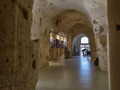 A long, arched interior corridor carved directly into light-colored tufa stone, featuring a modern smooth floor, track lighting, and framed photographs displayed on the left wall, with an arched doorway at the far end leading to daylight.