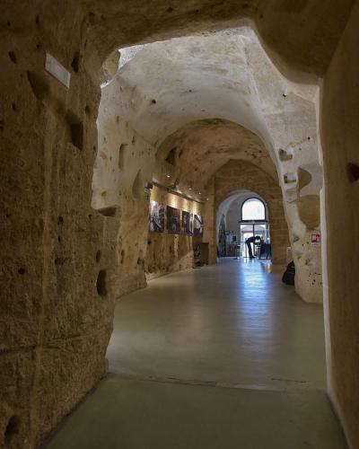 A long, arched interior corridor carved directly into light-colored tufa stone, featuring a modern smooth floor, track lighting, and framed photographs displayed on the left wall, with an arched doorway at the far end leading to daylight.