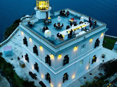 A high-angle evening shot of a white, multi-story lighthouse building perched on a cliff edge overlooking the sea, with its lantern room illuminated and its rooftop terrace set up as an outdoor restaurant or bar with patrons dining.