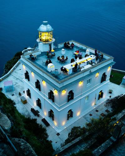 A high-angle evening shot of a white, multi-story lighthouse building perched on a cliff edge overlooking the sea, with its lantern room illuminated and its rooftop terrace set up as an outdoor restaurant or bar with patrons dining.