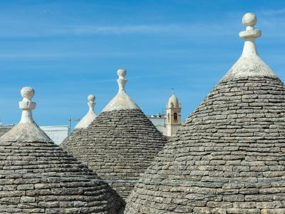 The image shows the conical stone roofs of several trulli with decorative pinnacles, set against a blue sky with a church bell tower visible in the background.