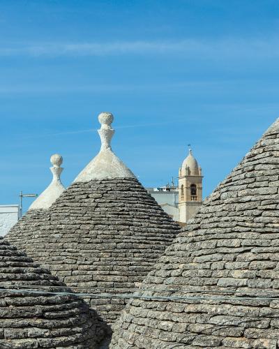 The image shows the conical stone roofs of several trulli with decorative pinnacles, set against a blue sky with a church bell tower visible in the background.