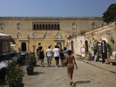 The image shows a sunlit courtyard of a historic stone building with Gothic-arched windows and a crenellated roofline, featuring an outdoor café on the left with a canopy labeled "CASTELLO" and tourists walking along a cobblestone path.