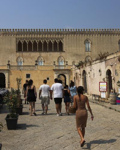 The image shows a sunlit courtyard of a historic stone building with Gothic-arched windows and a crenellated roofline, featuring an outdoor café on the left with a canopy labeled "CASTELLO" and tourists walking along a cobblestone path.