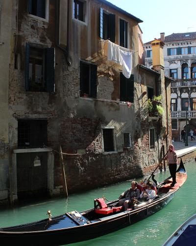 A close-up view of the bow of a black gondola moored against a weathered, plastered canal wall with two windows above.