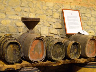 The image shows a display of old wooden wine barrels and a large wooden funnel arranged on a wooden shelf against a rough stone wall, with an informational placard in the background.