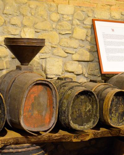 The image shows a display of old wooden wine barrels and a large wooden funnel arranged on a wooden shelf against a rough stone wall, with an informational placard in the background.