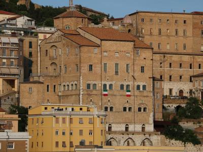 A large, historic brick building with arched windows, two Italian flags hanging from its facade, and a terracotta roof, situated on a hillside among other buildings in an urban Italian setting.