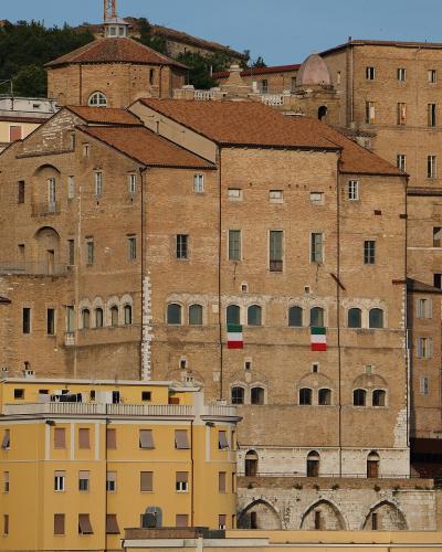 A large, historic brick building with arched windows, two Italian flags hanging from its facade, and a terracotta roof, situated on a hillside among other buildings in an urban Italian setting.