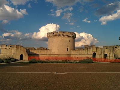 A large medieval stone fortress with a prominent central round tower and crenellated walls, set against a blue sky with clouds, featuring an orange safety fence in the foreground.