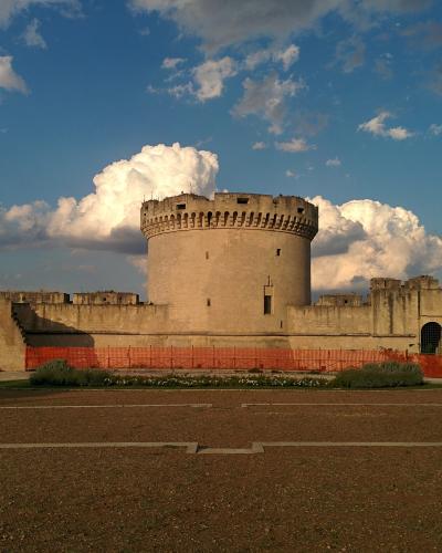 A large medieval stone fortress with a prominent central round tower and crenellated walls, set against a blue sky with clouds, featuring an orange safety fence in the foreground.